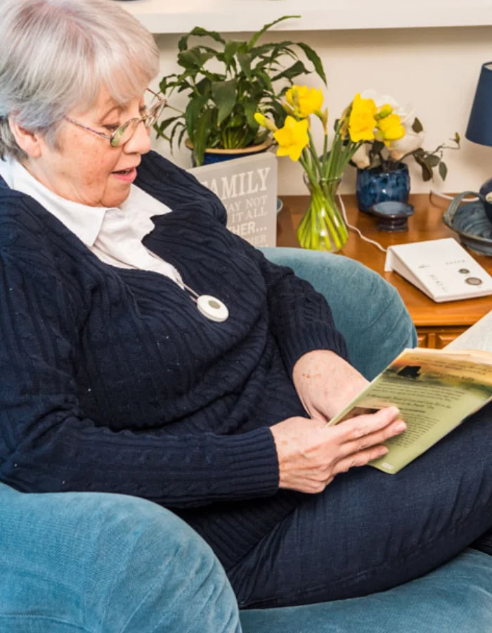 Elderly Lady at reading a book, wearing a telecare pendant and next to a home hub